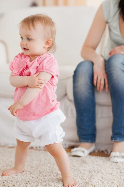 Baby standing on a carpet while her mother is sitting on a sofa — Stock Photo, Image