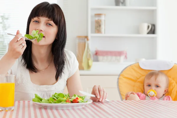 Attraktive brünette Frau isst einen Salat neben ihrem Baby, während — Stockfoto