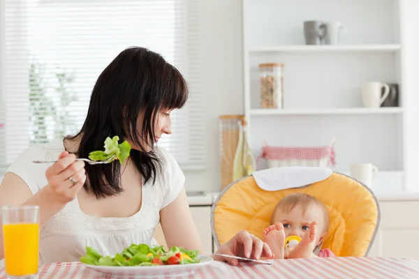 Beautiful brunette woman eating a salad next to her baby while s — Stock Photo, Image