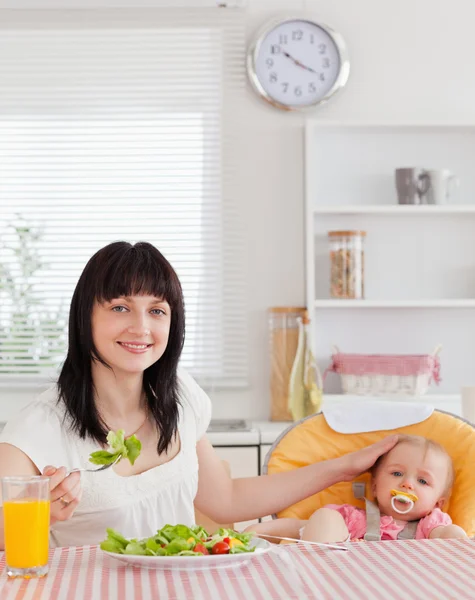 Mulher morena bonita comendo uma salada ao lado de seu bebê enquanto sentada — Fotografia de Stock