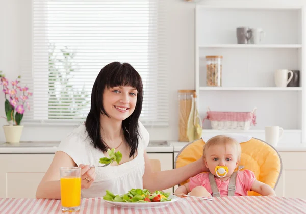 Mulher morena encantadora comendo uma salada ao lado de seu bebê enquanto si — Fotografia de Stock