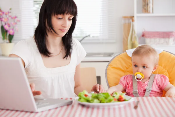 Gorgeous brunette woman eating a salad next to her baby while re — Stock Photo, Image