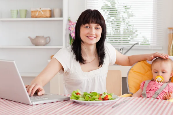 Attractive brunette woman eating a salad next to her baby while — Stock Photo, Image