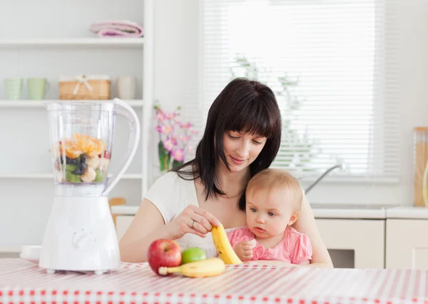 Cute brunette woman showing a banana to her baby while sitting — Stock Photo, Image