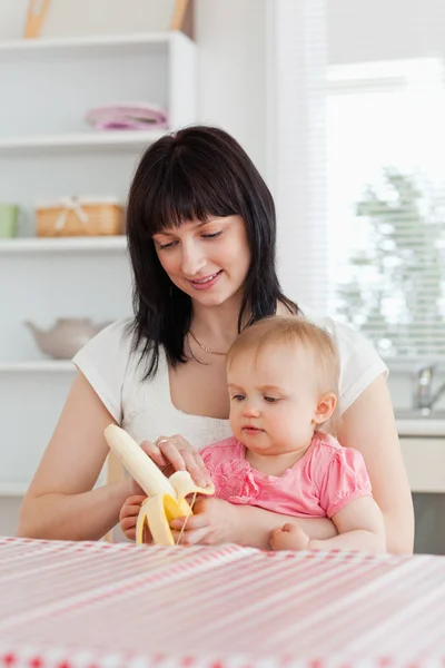 Beautiful brunette woman pealing a banana while holding her baby — Stock Photo, Image