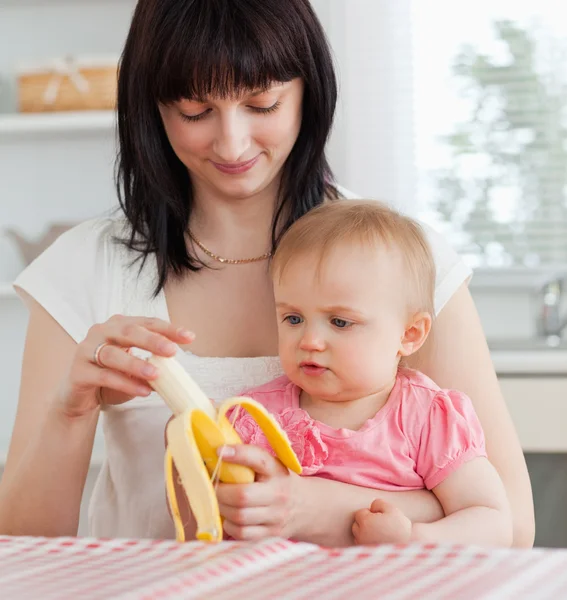 Förtjusande brunett kvinna pealing en banan medan du håller hennes baby — Stockfoto