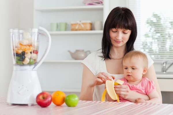 Gorgeous brunette woman pealing a banana while holding her baby — Stock Photo, Image