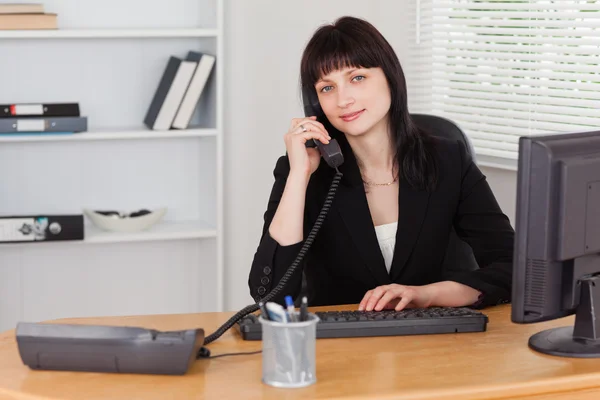 Hübsche brünette Frau am Telefon, während sie an einem Computer arbeitet — Stockfoto