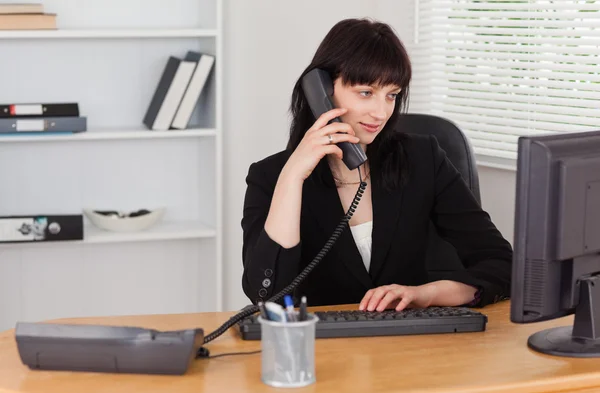 Attraktive brünette Frau am Telefon, während sie an einem Computer arbeitet — Stockfoto