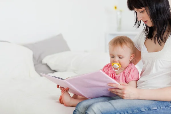 Pretty brunette woman showing a book to her baby while sitting o — Stock Photo, Image