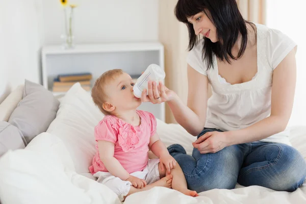 Uroczy brunetka kobieta karmienie za pomocą butelki swojego dziecka na łóżku — Zdjęcie stockowe