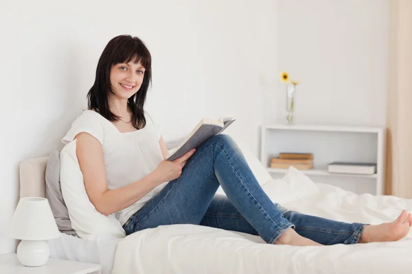 Attractive brunette woman reading a book while sitting on a bed — Stock Photo, Image
