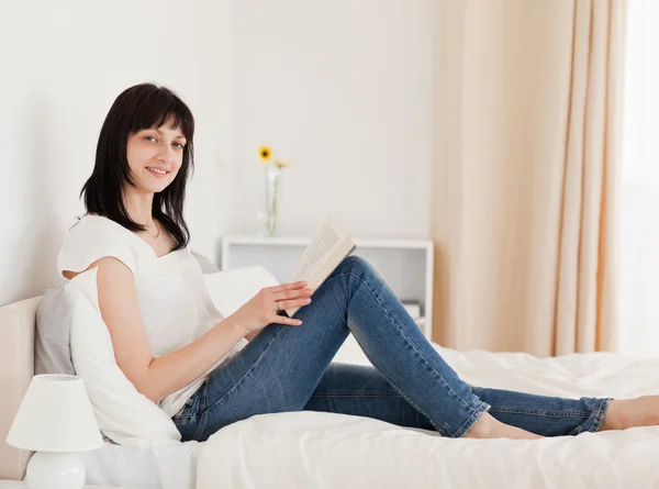 Gorgeous brunette woman reading a book while sitting on a bed — Stock Photo, Image