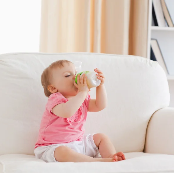 Szép szőke baba üvegből történő táplálás, a kanapén ülve — Stock Fotó