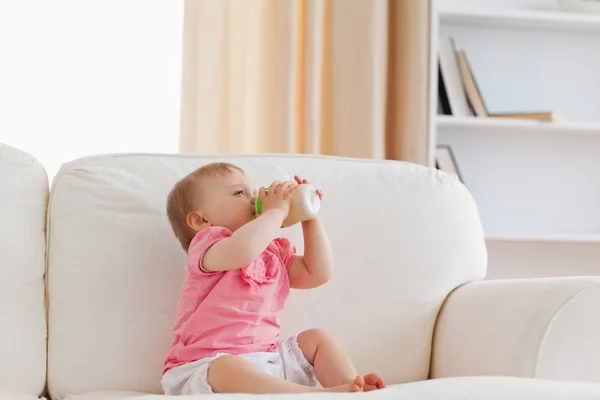Ładny blond dziecko karmienie za pomocą butelki, siedząc na kanapie — Zdjęcie stockowe