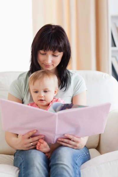 Mujer bonita sosteniendo a su bebé y un libro en sus brazos mientras se sienta —  Fotos de Stock
