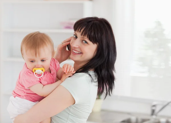 Gut aussehende Frau am Telefon, während sie ihr Baby im Arm hält — Stockfoto