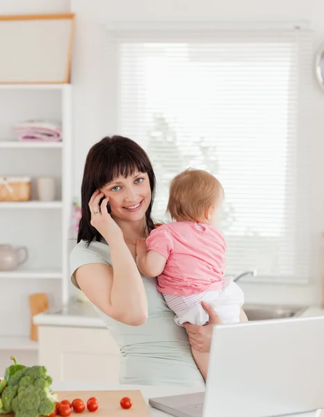 Schöne brünette Frau am Telefon, während sie ihr Baby in der Hand hält — Stockfoto