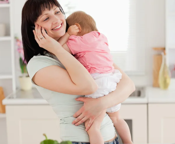 Schöne brünette Frau am Telefon, während sie ihr Baby in der Hand hält — Stockfoto