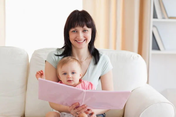 Mujer guapa sosteniendo a su bebé y un libro en sus brazos mientras — Foto de Stock