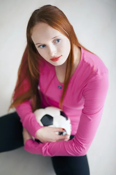 吸引人的金发女孩拿着一个足球球和坐在地板上 — 图库照片