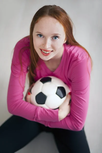吸引人的金发女孩拿着一个足球球和坐在地板上 — 图库照片