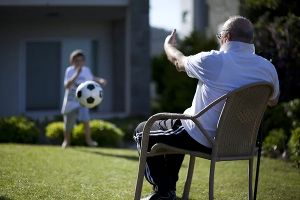 Farfar sitter på stol och hans sonson spela fotboll — Stockfoto