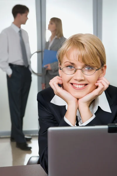 Молодая привлекательная офисная работница смотрит на камеру, трогающую ноутбук на фоне своих коллег — стоковое фото