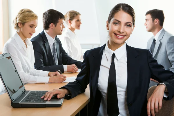 Young attractive office worker is looking at camera touching the laptop on the background of her colleagues — Stock Photo, Image