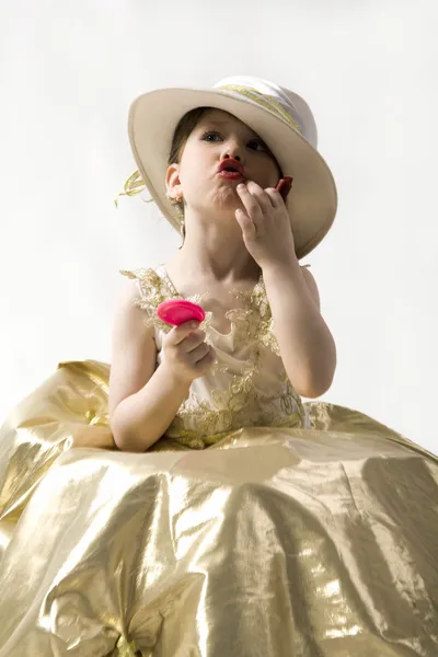 Sweet brunette little girl in light goldish dress and white hat making up her lips — Stock Photo, Image