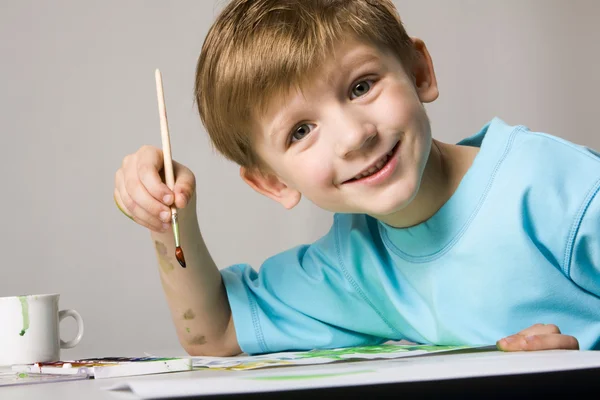 Lindo niño sonriente está sentado en la mesa y sosteniendo el pincel en su mano —  Fotos de Stock