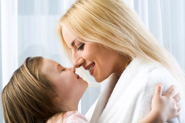Niña rubia abrazando a su sonriente madre alcanzando a besarla —  Fotos de Stock