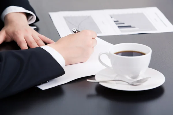 Mão de mulher de negócios escrevendo algo no papel com xícara de café e documento por perto — Fotografia de Stock